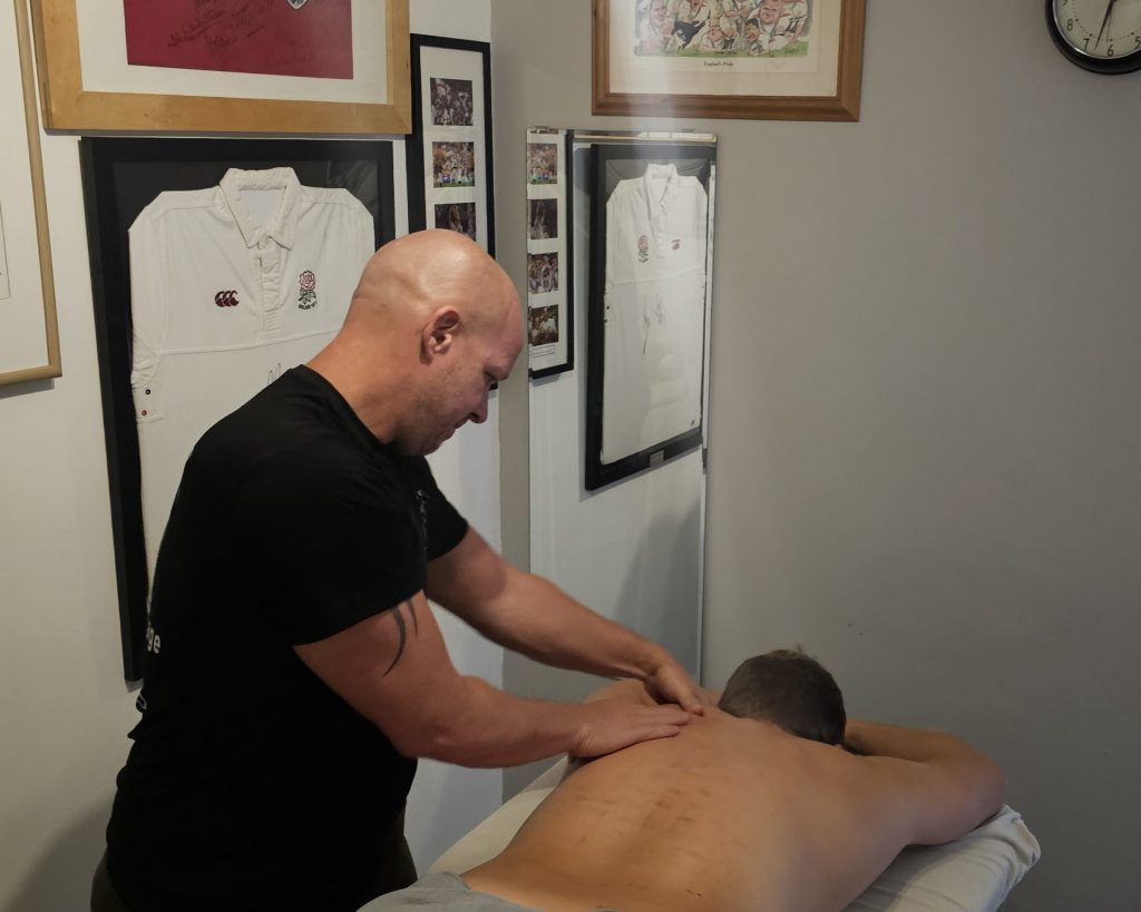 Sports massage therapist Matt Edwards performing a back massage on a client, with framed sports memorabilia (including England rugby shirts) in the background.