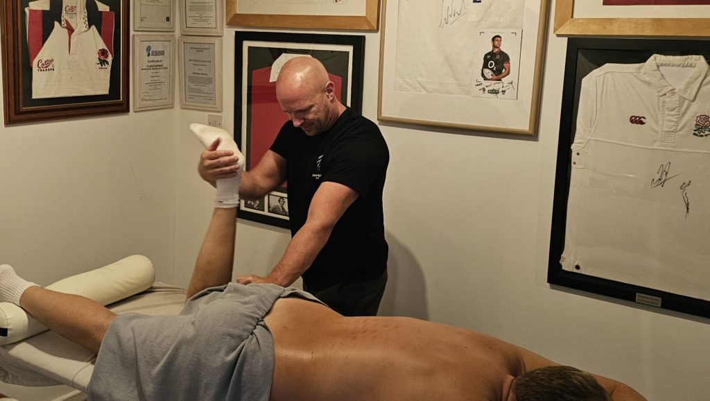 Sports massage therapist Matt Edwards performing a passive hamstring stretch and glute mobilization on a client in a treatment room decorated with framed England rugby shirts.